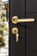 A close-up of a modern black front door with a brushed gold lever handle and matching lockset, with keys inserted and an outdoor garden background visible through the open door.